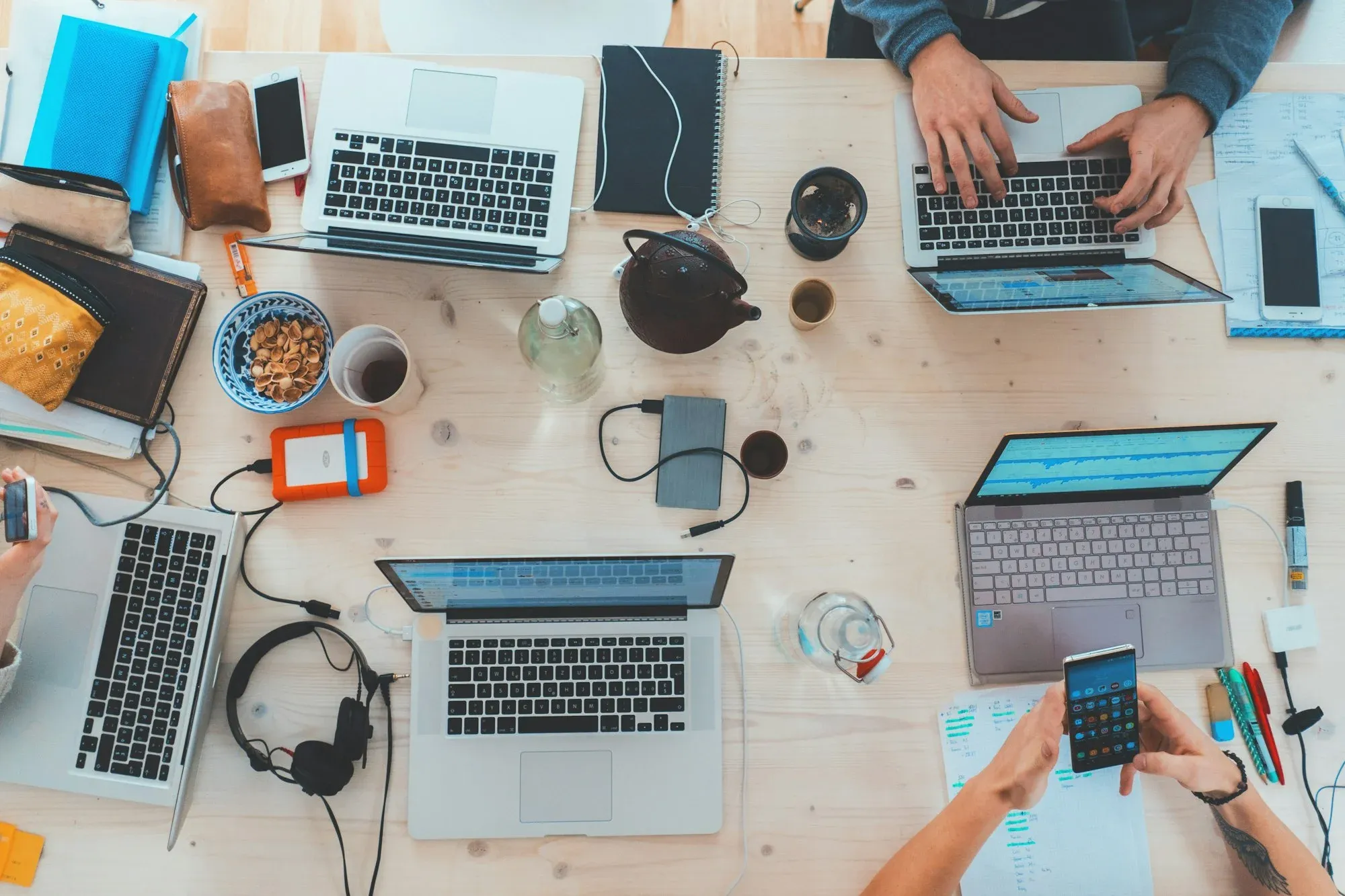A desk covered with several open laptops, notebooks, cables and phones being used by different people at once